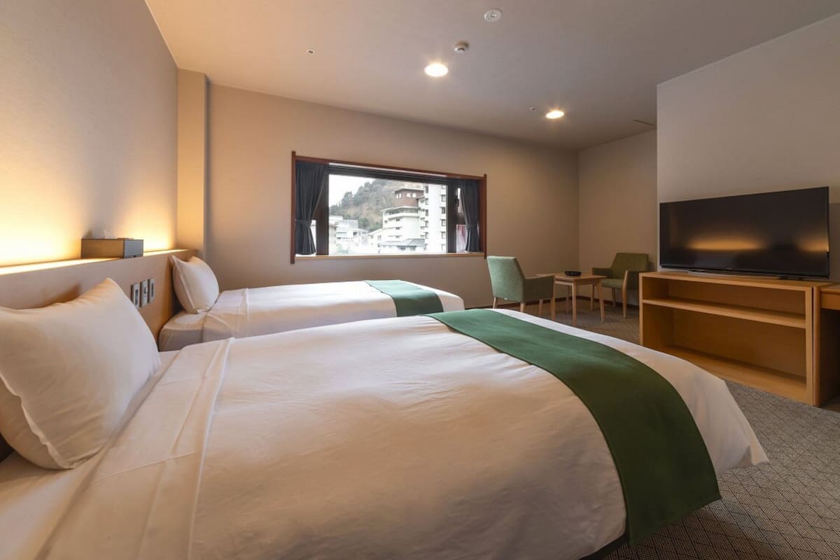 A hotel room with two beds, a television, a desk, and a window with a view of buildings.