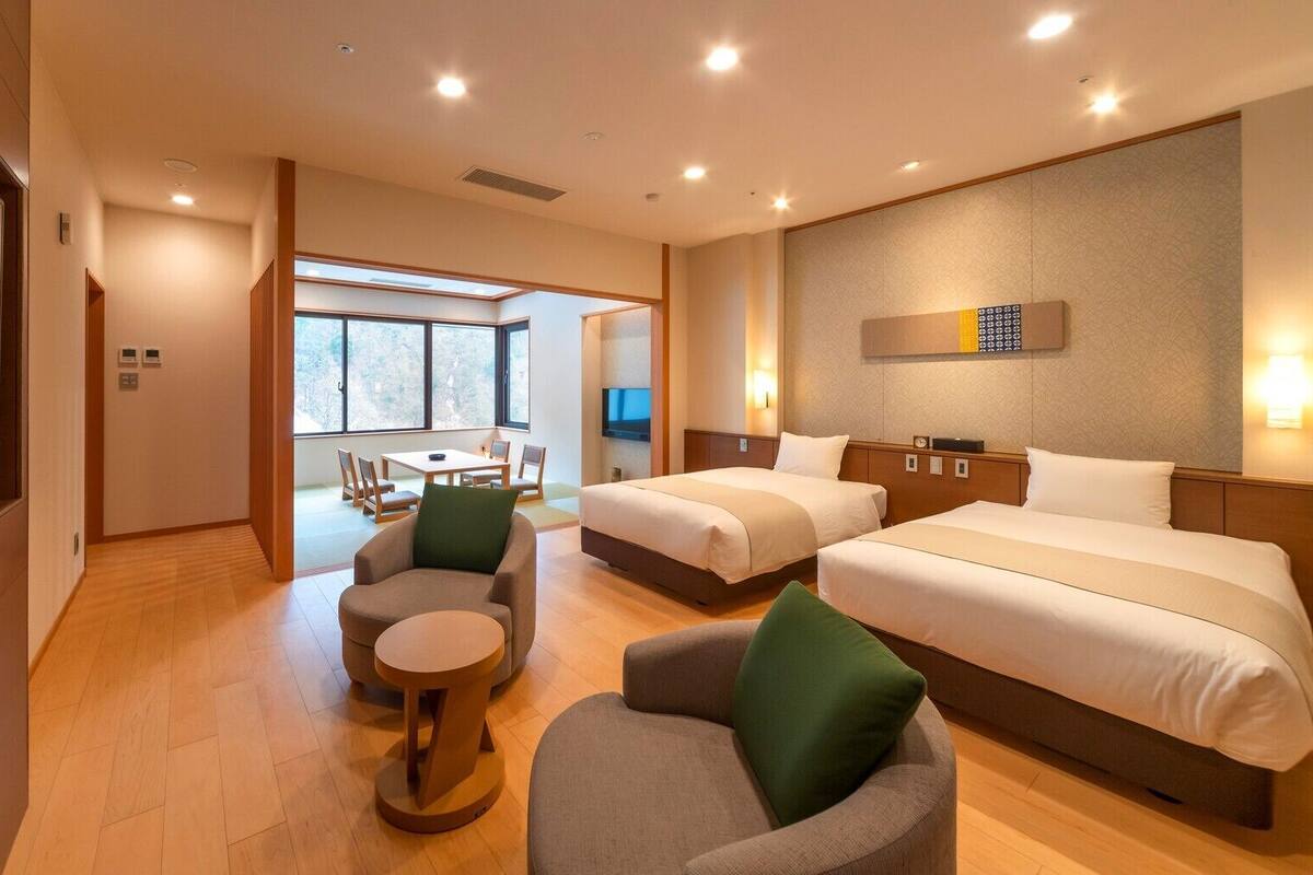 A hotel room with two beds, a TV, a dining area with a table and chairs, and a large window.