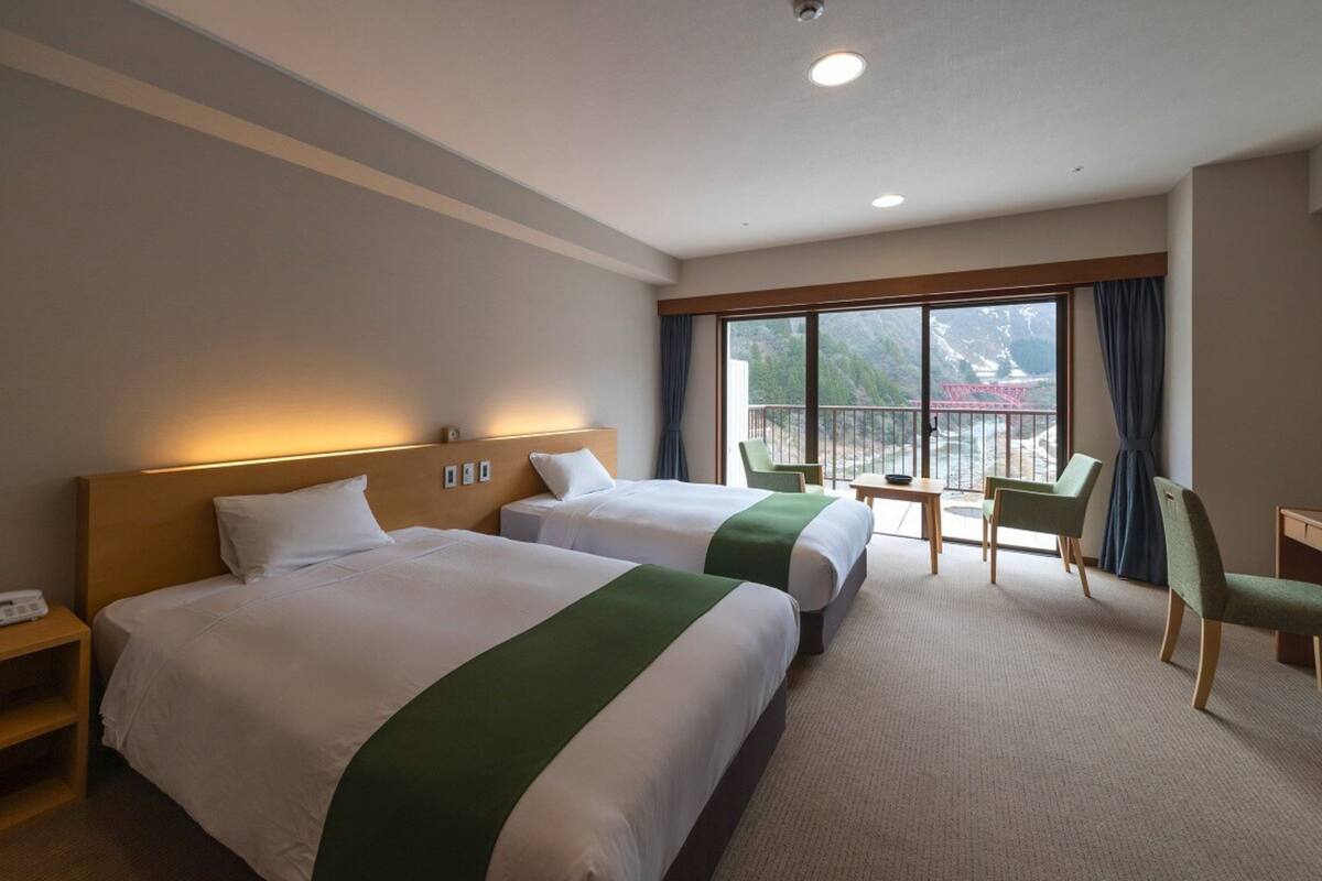 A hotel room with two beds, a desk, chairs, and a balcony with a view.