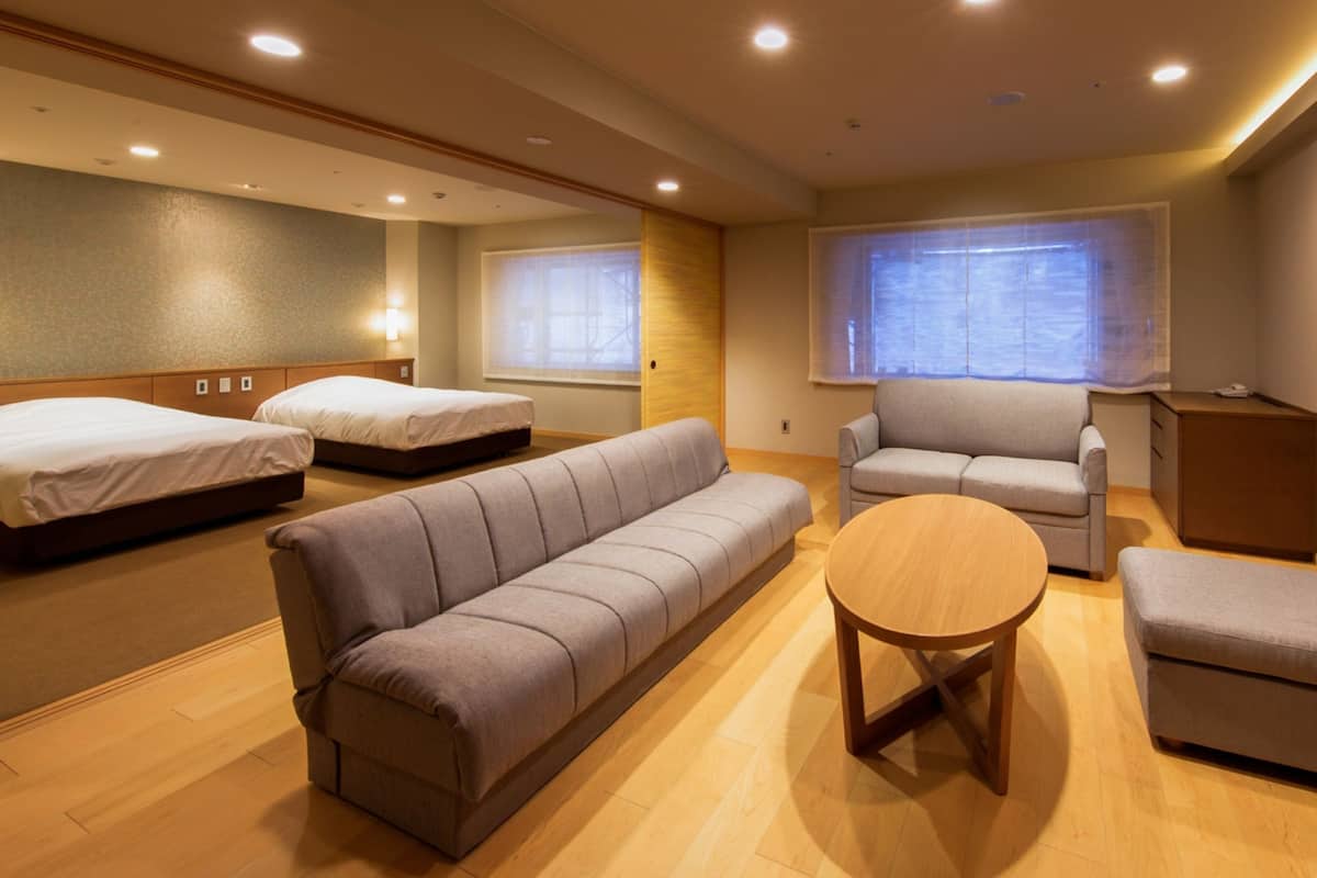 A hotel room with two single beds, a grey sofa, a round wooden table, and a grey armchair.