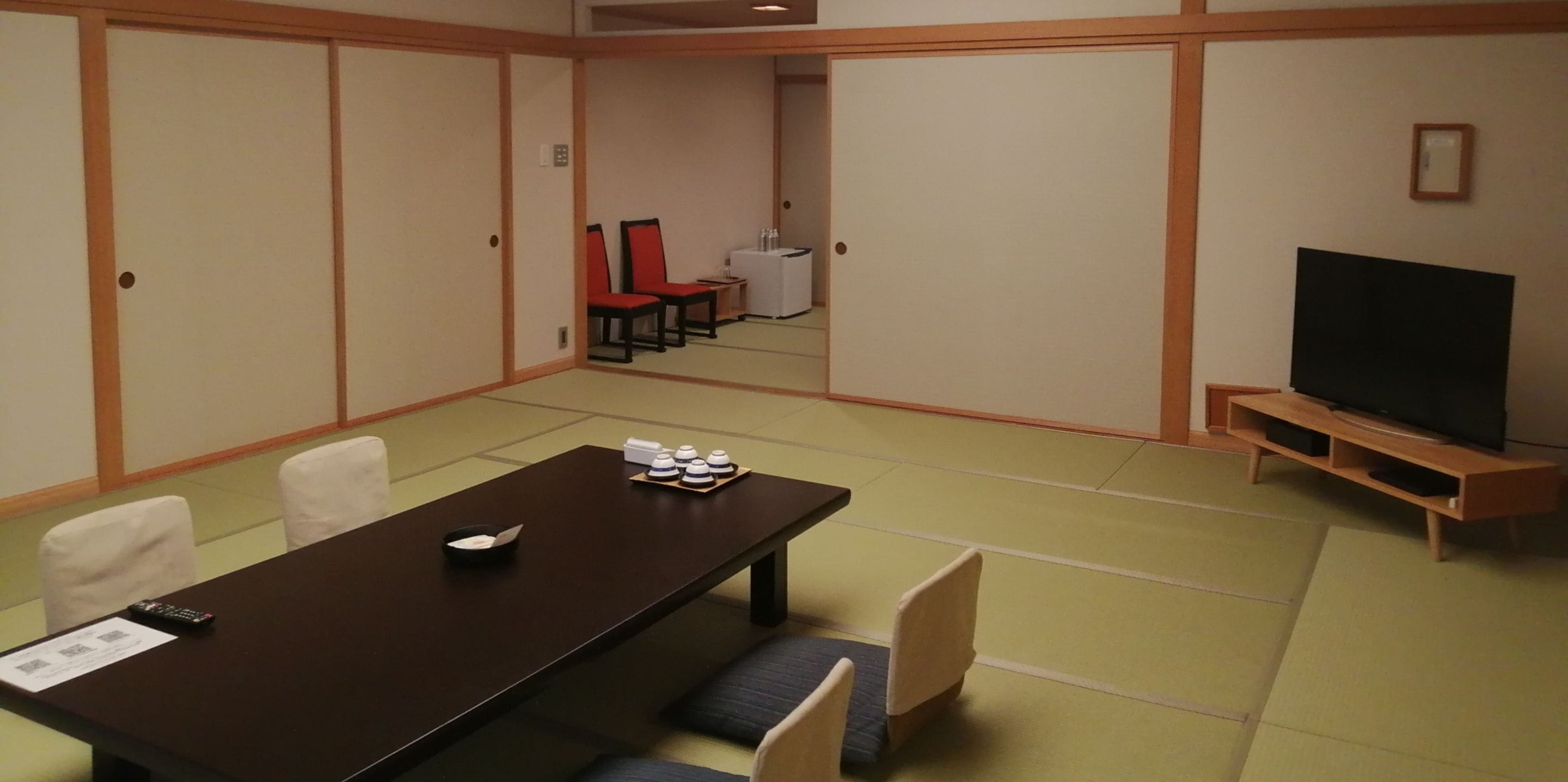 Une salle de conférence avec une grande table en bois foncé, des chaises blanches, un téléviseur posé sur un meuble en bois et une petite table sur laquelle se trouvent des tasses et une télécommande.