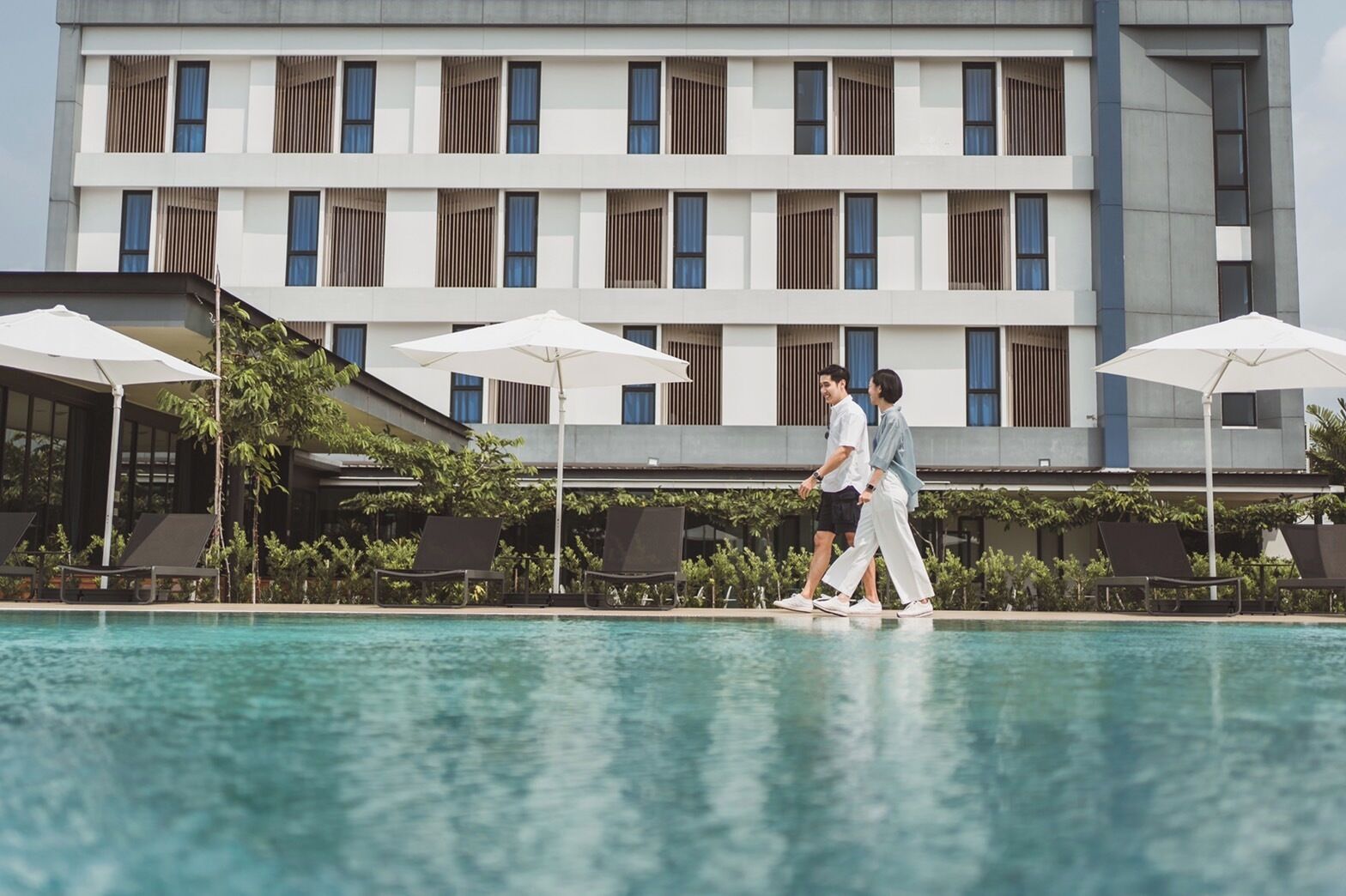 Un hotel moderno con área de piscina, que presenta un edificio grande con múltiples ventanas y sombrillas exteriores.