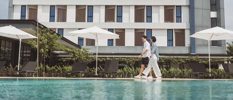 Un hotel moderno con área de piscina, que presenta un edificio grande con múltiples ventanas y sombrillas exteriores.