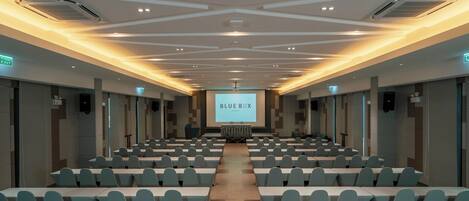 Un salón de conferencias con filas de mesas y sillas, un escenario con una pantalla que muestra “BLUE BOX” y luces en el techo.