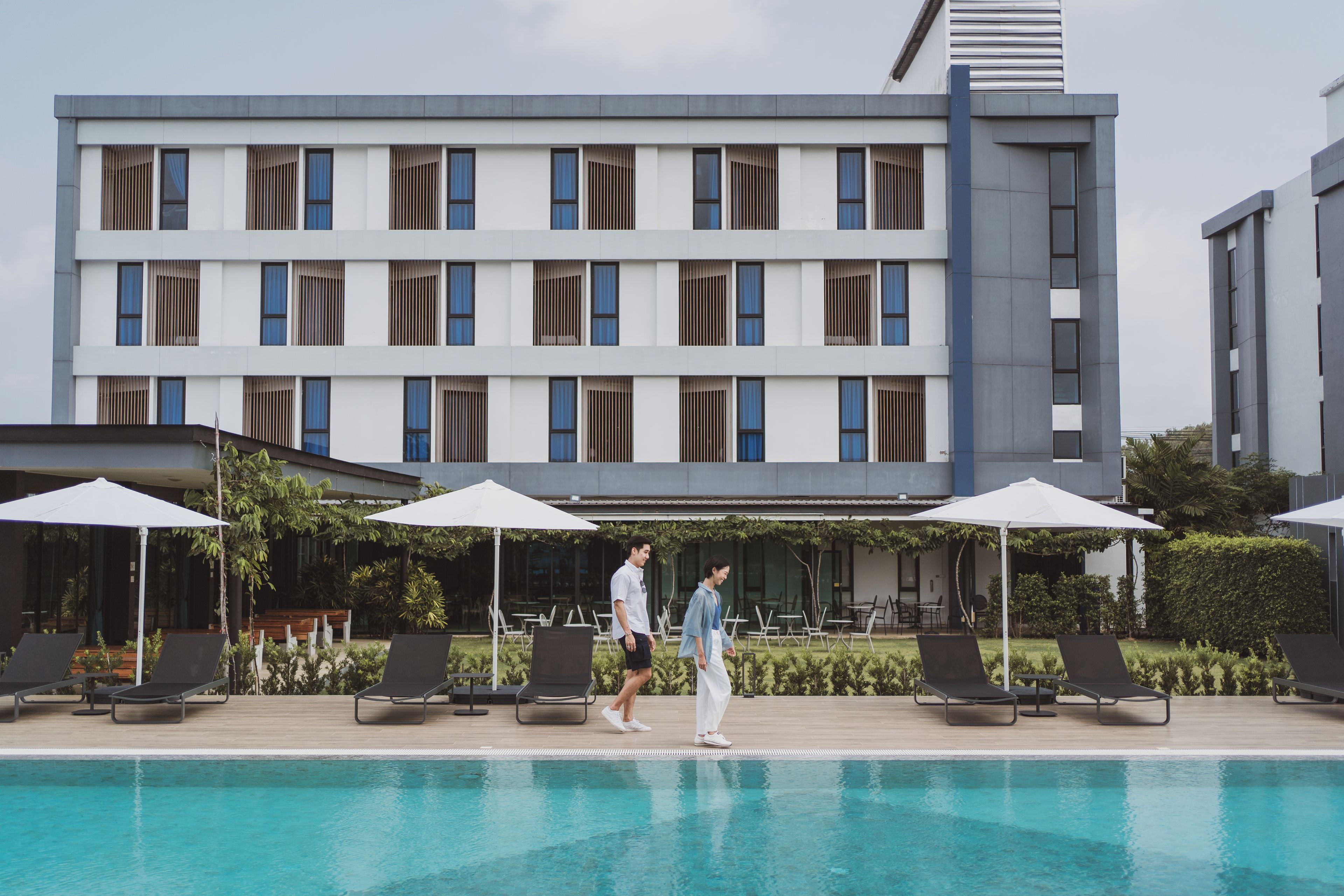 Un hotel moderno con piscina, zona de estar exterior y un edificio de varios pisos con grandes ventanales.