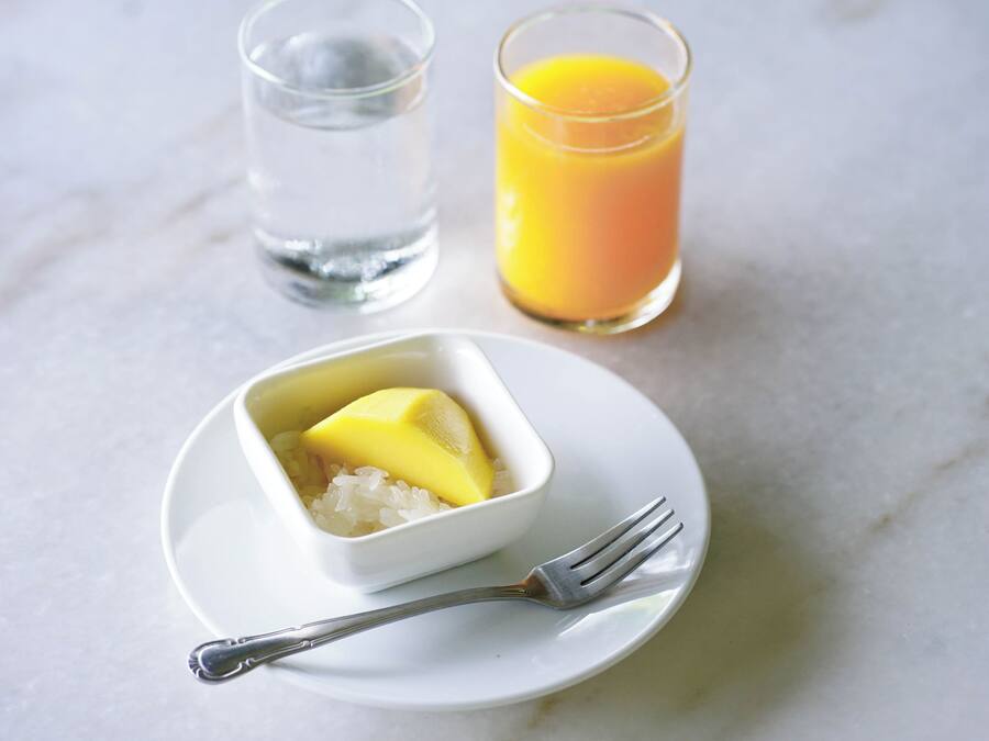 Een wit bord met een kleine schaal met een geel toetje, een glas sinaasappelsap en een glas water op een marmeren oppervlak.