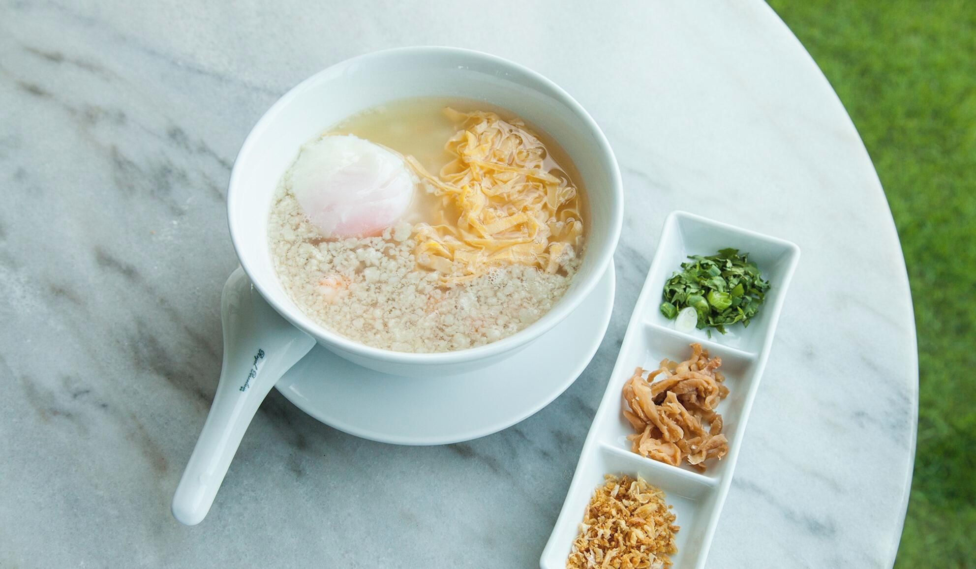 A bowl of soup with a poached egg, noodles, and a side dish of greens and fried items on a marble table.