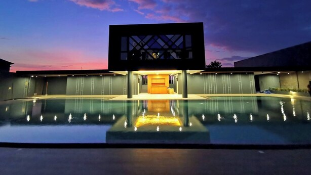 Edificio moderno con piscina en primer plano, un área techada con escalones que conducen a una puerta y un cielo al atardecer en el fondo.
