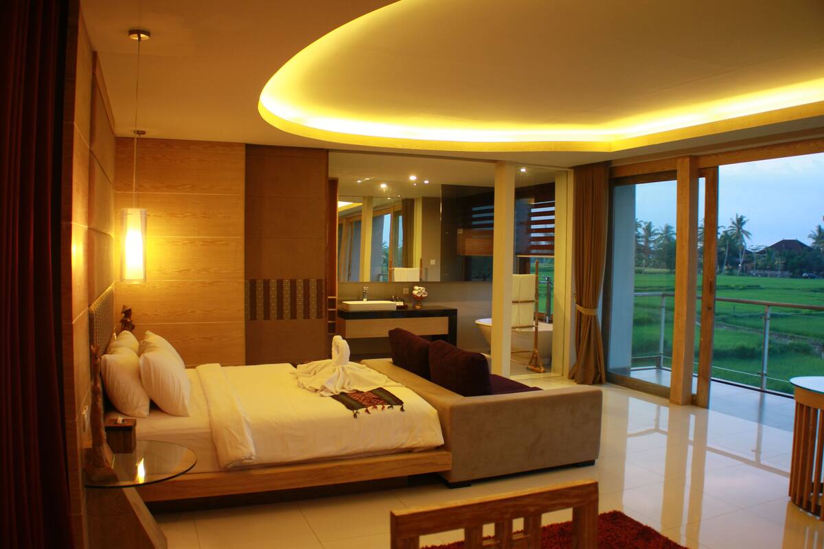 A modern hotel room with a large bed, a sitting area, a bathroom, and a view of the outdoors.
