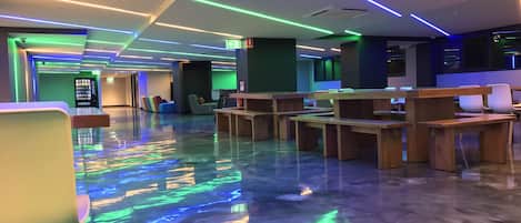 A modern lobby with a polished floor reflecting colorful LED lights, wooden seating areas, and a vending machine.