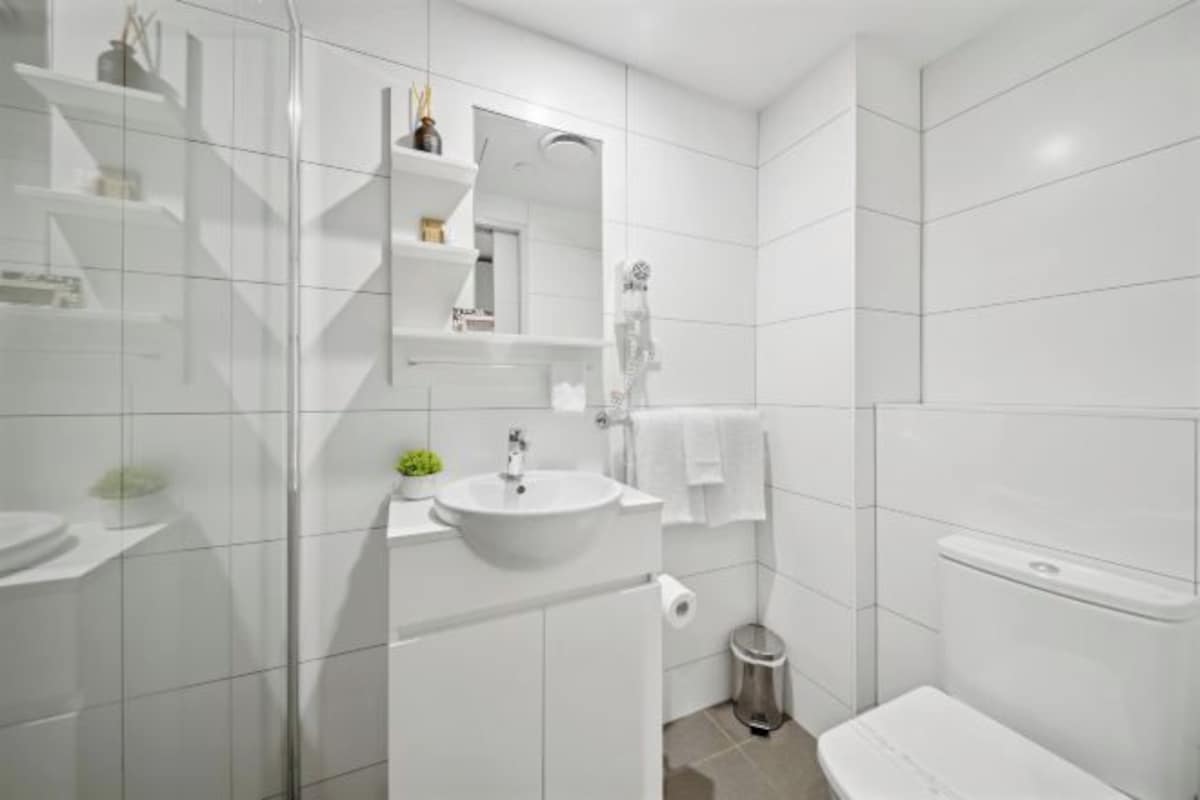 Un baño moderno con un lavamanos blanco, espejo e inodoro. Hay repisas sobre el lavamanos y un cesto de basura junto al inodoro.