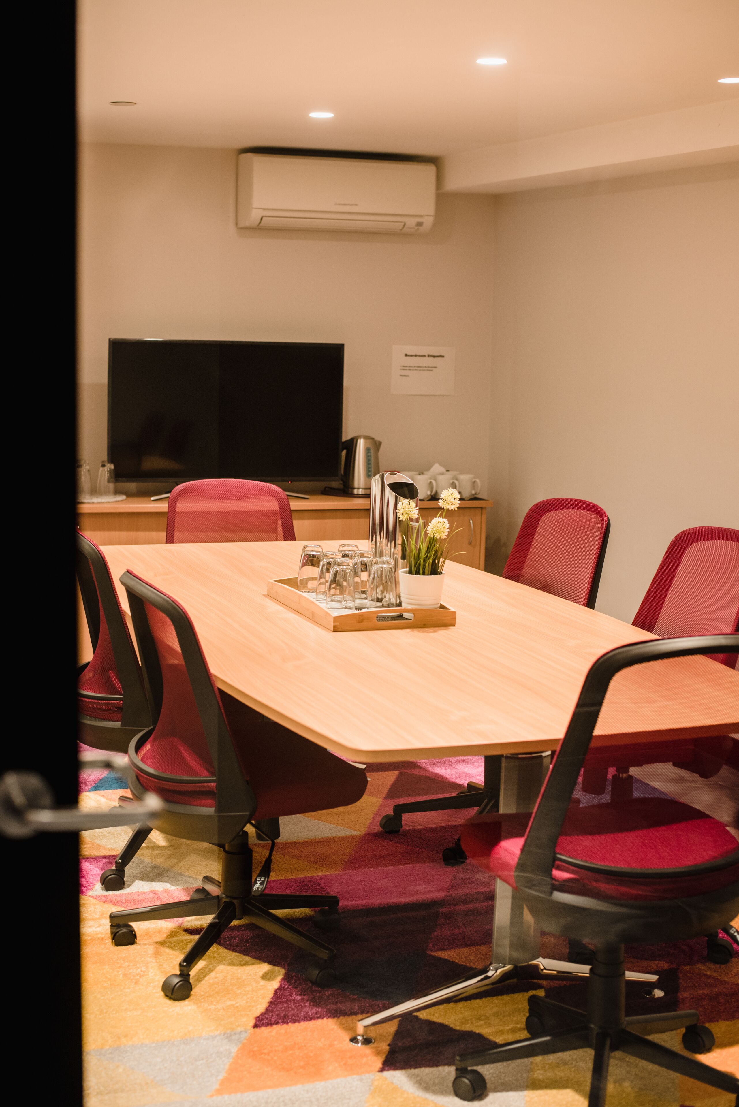 A conference room with a long table, red chairs, a flat-screen TV, and a coffee machine.