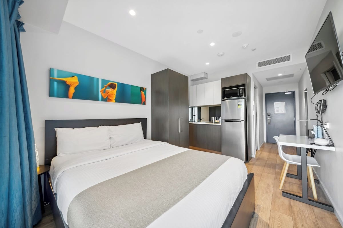 A modern hotel room with a large bed, a kitchenette, a desk with a chair, and a TV mounted on the wall.