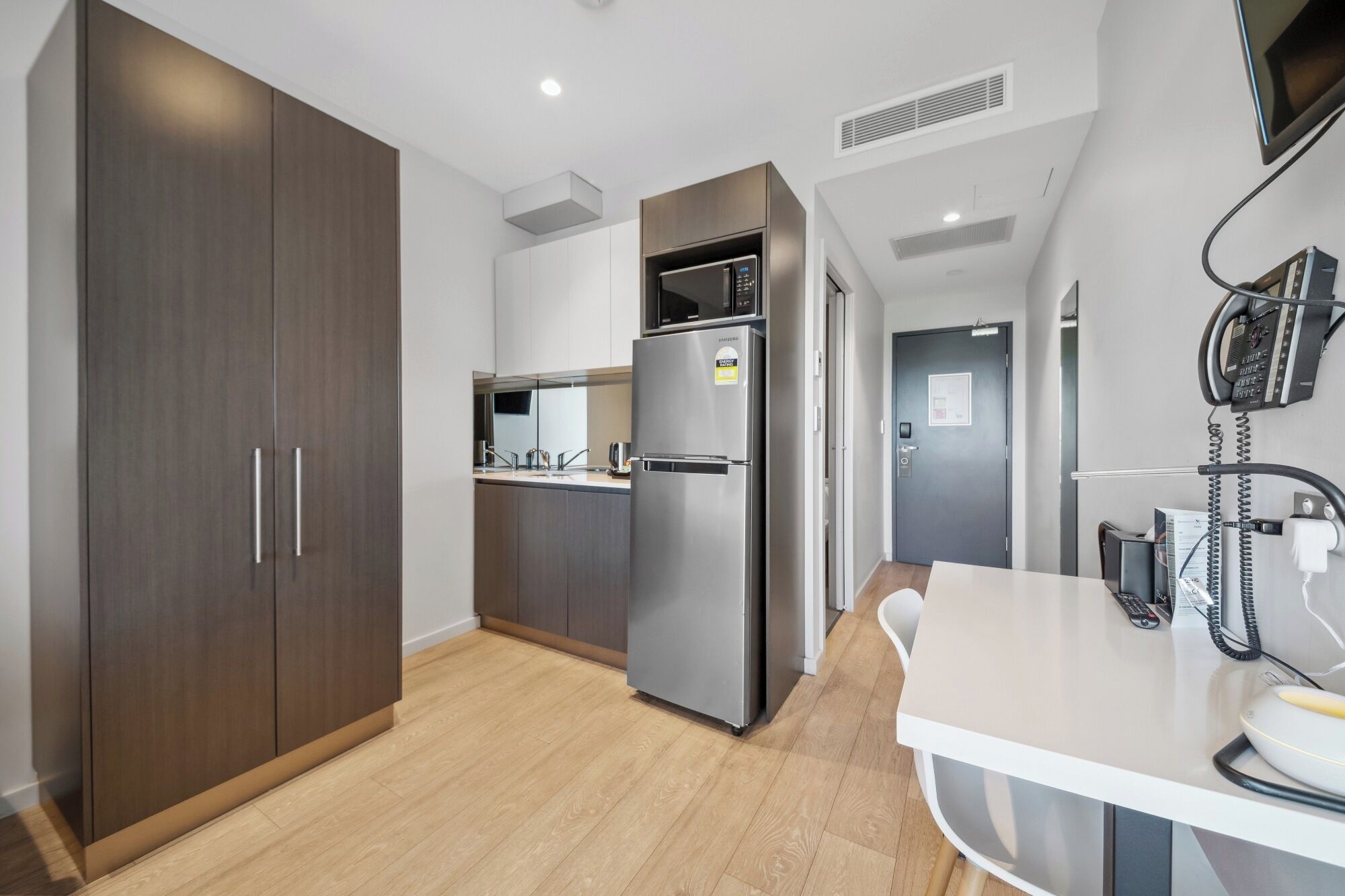 Una cocina moderna con armarios de madera oscura, un refrigerador de acero inoxidable y un microondas. Hay una encimera blanca con un teléfono y una taza.