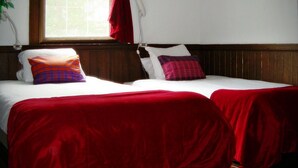 A room with two beds, each with a red bedspread and pillows, a window with red curtains, and a white lamp on a bedside table. - Australia House (Kutchan)
