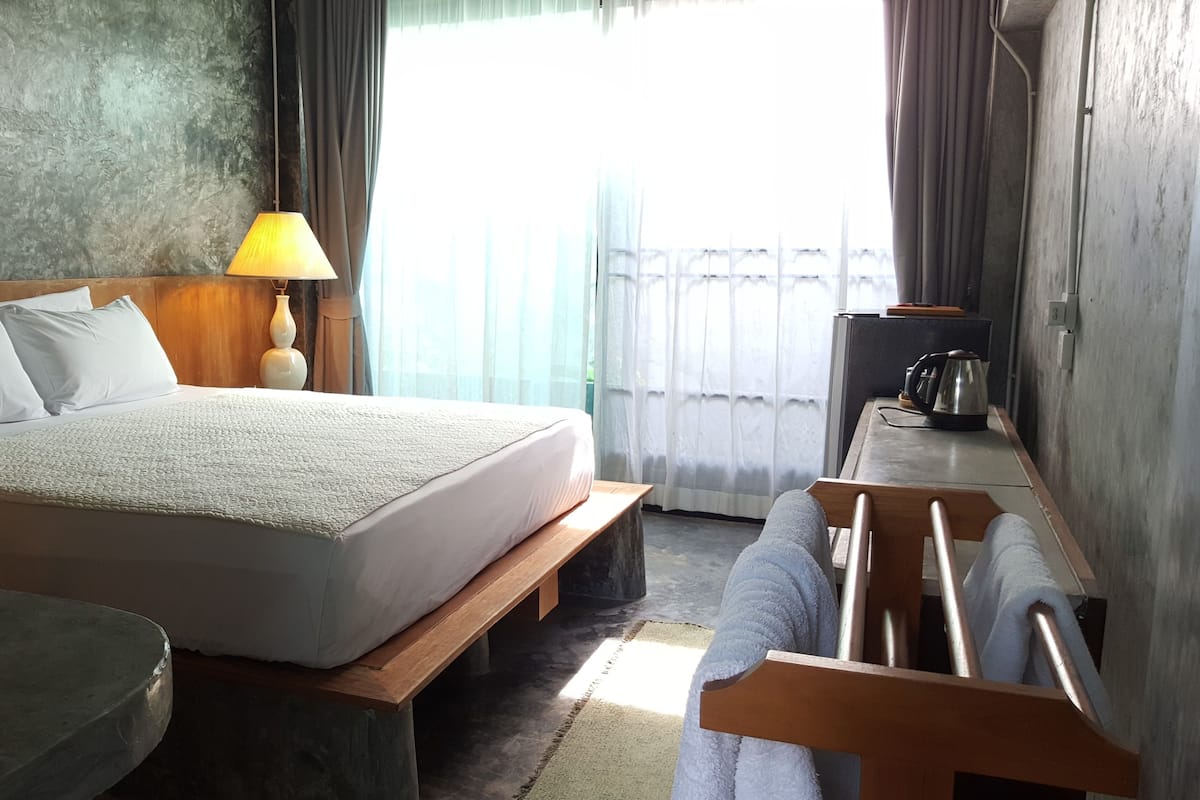 A hotel room with a bed, bedside lamp, wooden bench, and a view of the outside.