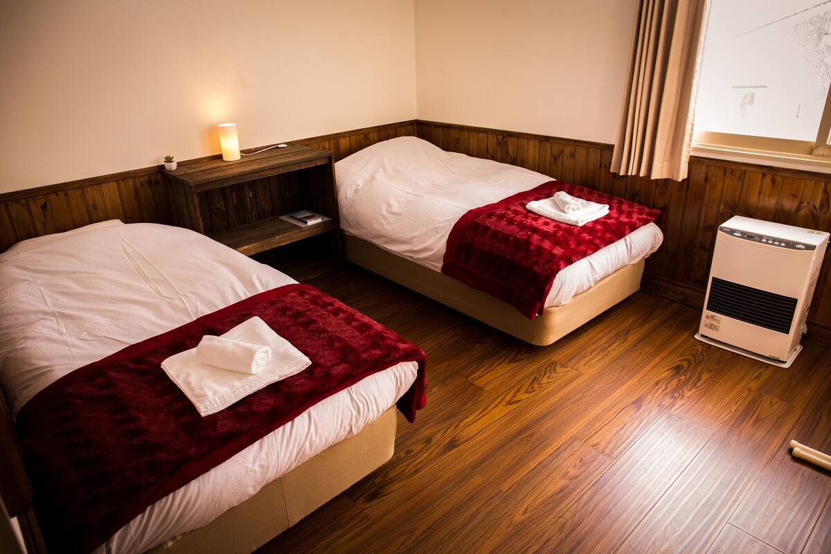 Dos camas individuales con mantas rojas y almohadas blancas, una mesita de noche con lámpara y un calefactor en el suelo.