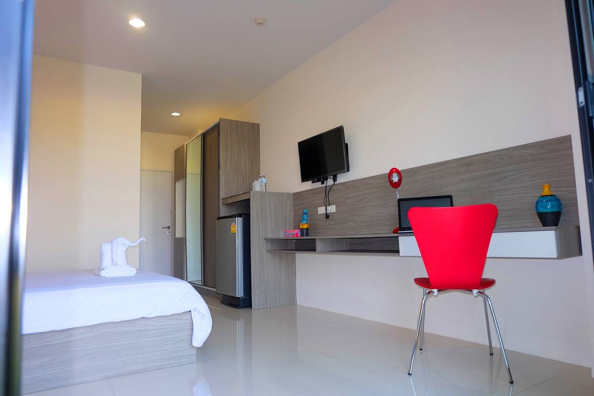 A modern hotel room with a bed, a red chair, a TV, and a minibar.