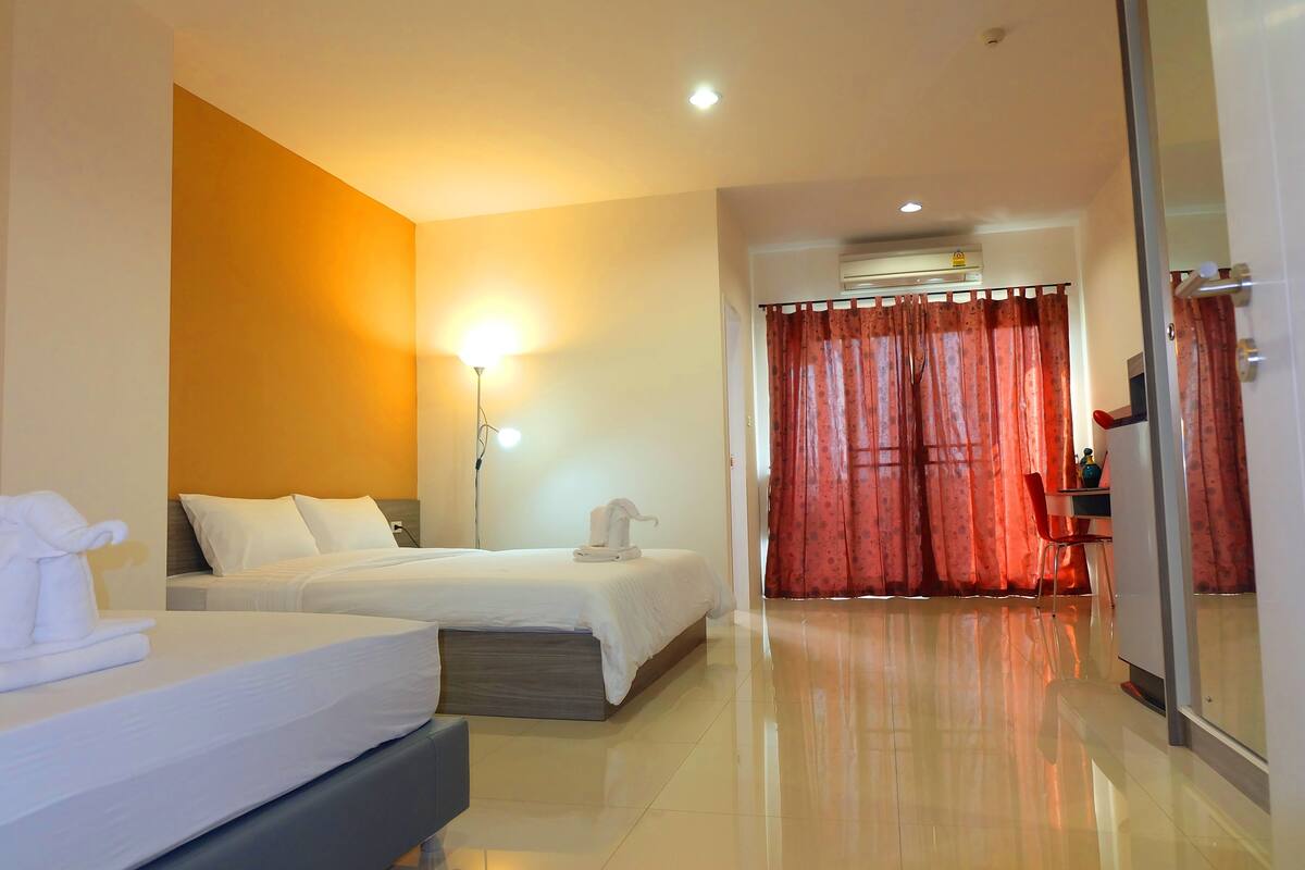 A hotel room with two beds, a bedside table, a lamp, a mirror, and a sliding door with red curtains.