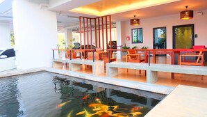 A modern indoor pool area with a koi pond, wooden seating, and a view of the outdoors. - The Pride (Sam Phran)