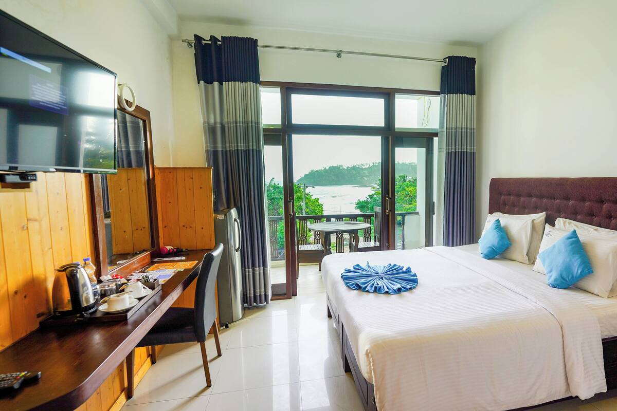 A hotel room with a bed, a desk with a computer, a TV, and a balcony with a view of the sea.