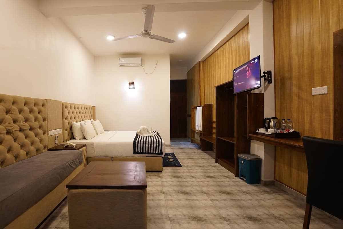 A hotel room with a large tufted headboard, a bed with white bedding, a small sofa, a wooden coffee table, a TV mounted on the wall, and a wooden wardrobe.
