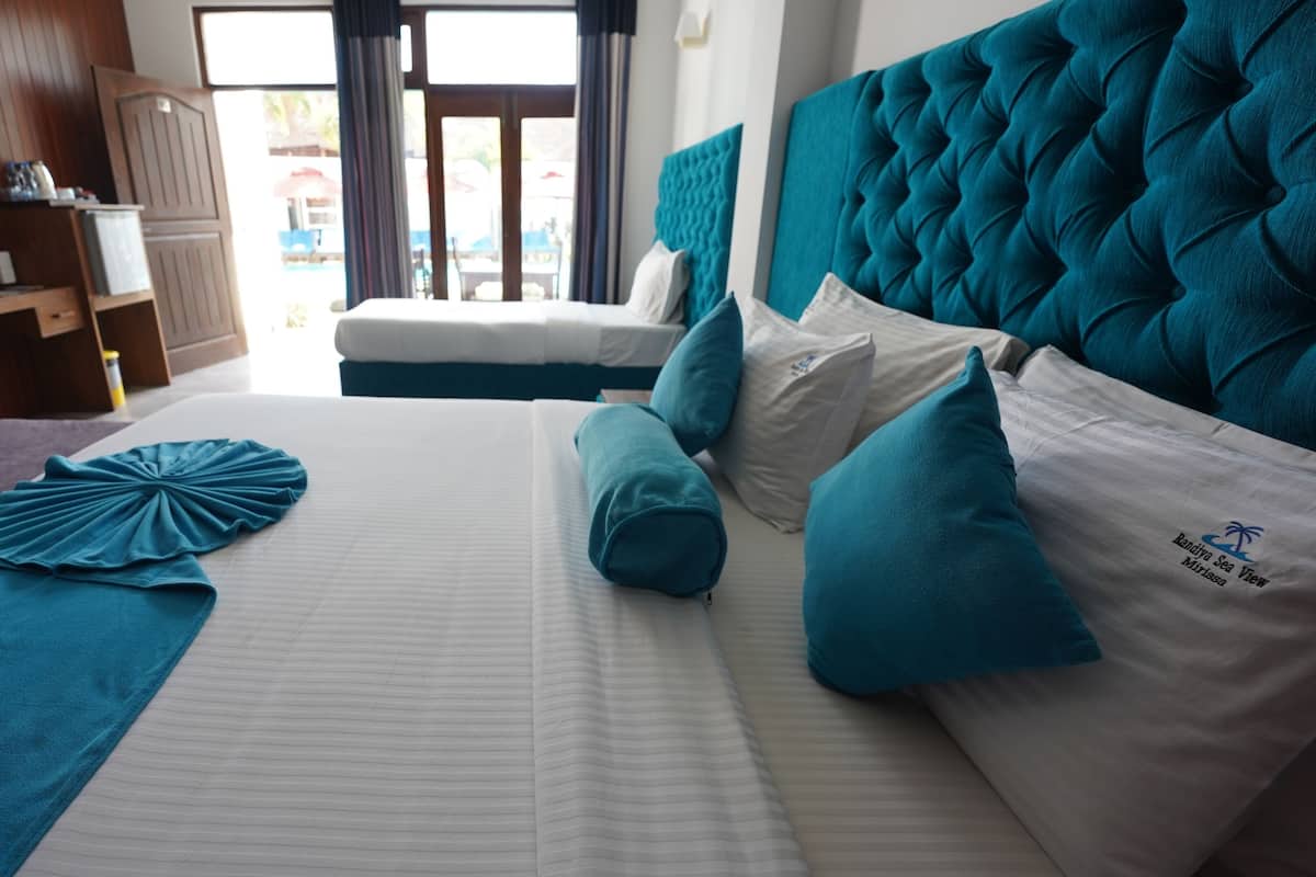 A hotel room with a large bed, teal and white bedding, a television on a wooden cabinet, and a view of the outside through a door.