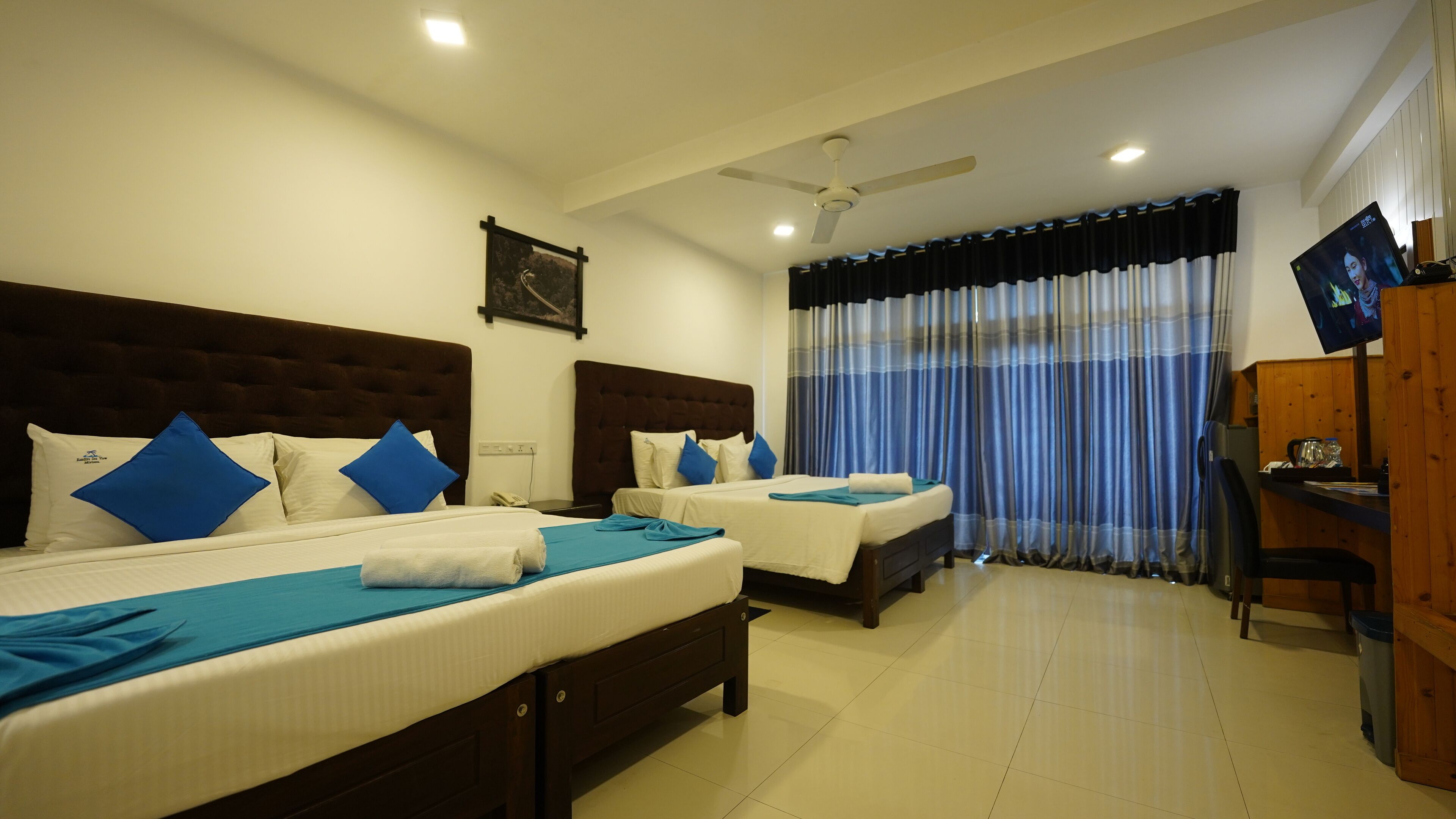 A hotel room with two beds, each with a headboard, a desk with a chair, a TV, and a window with curtains.