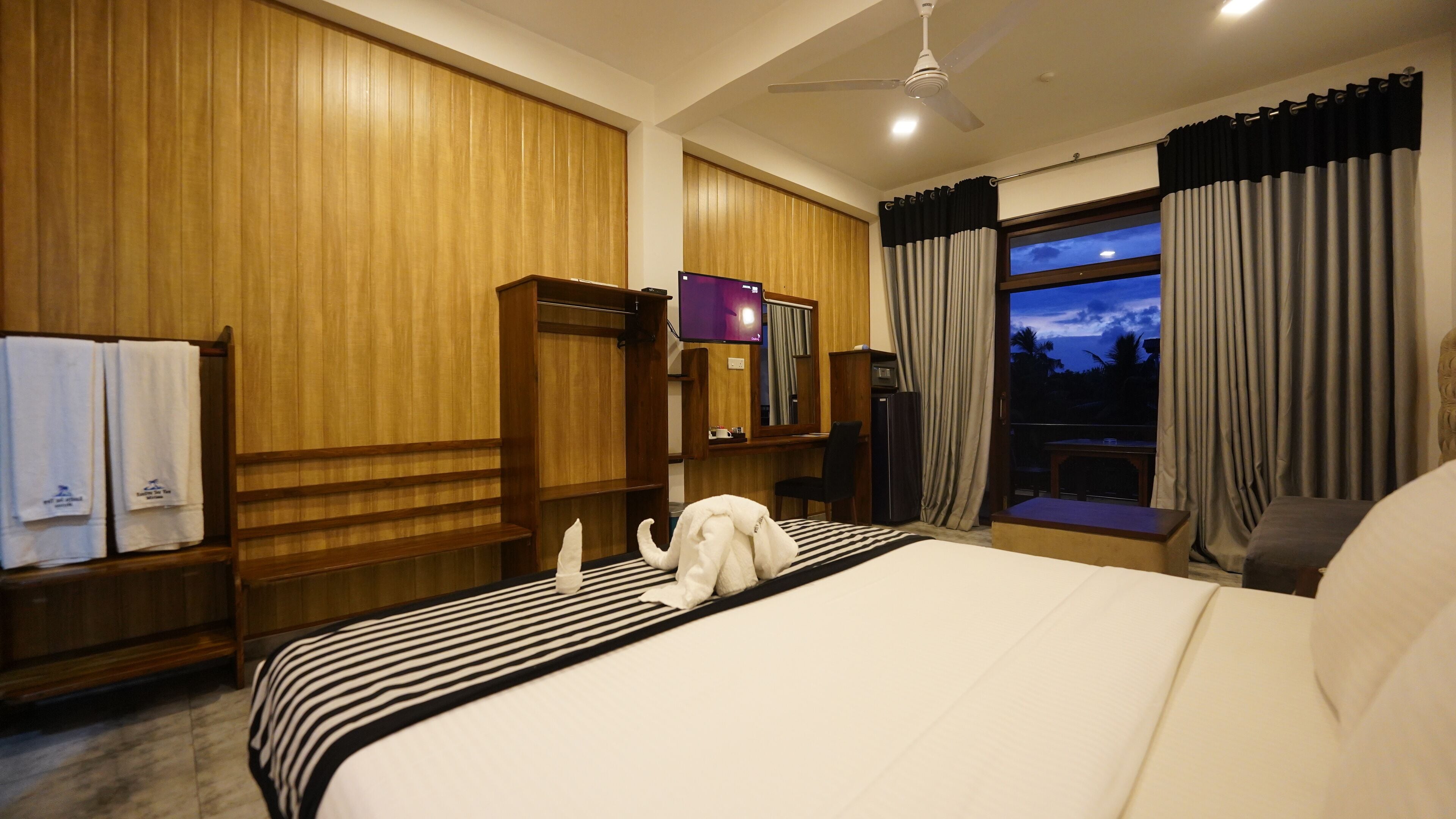 A hotel room with a large bed, wooden paneling, a television, a desk, and a view of the evening sky.
