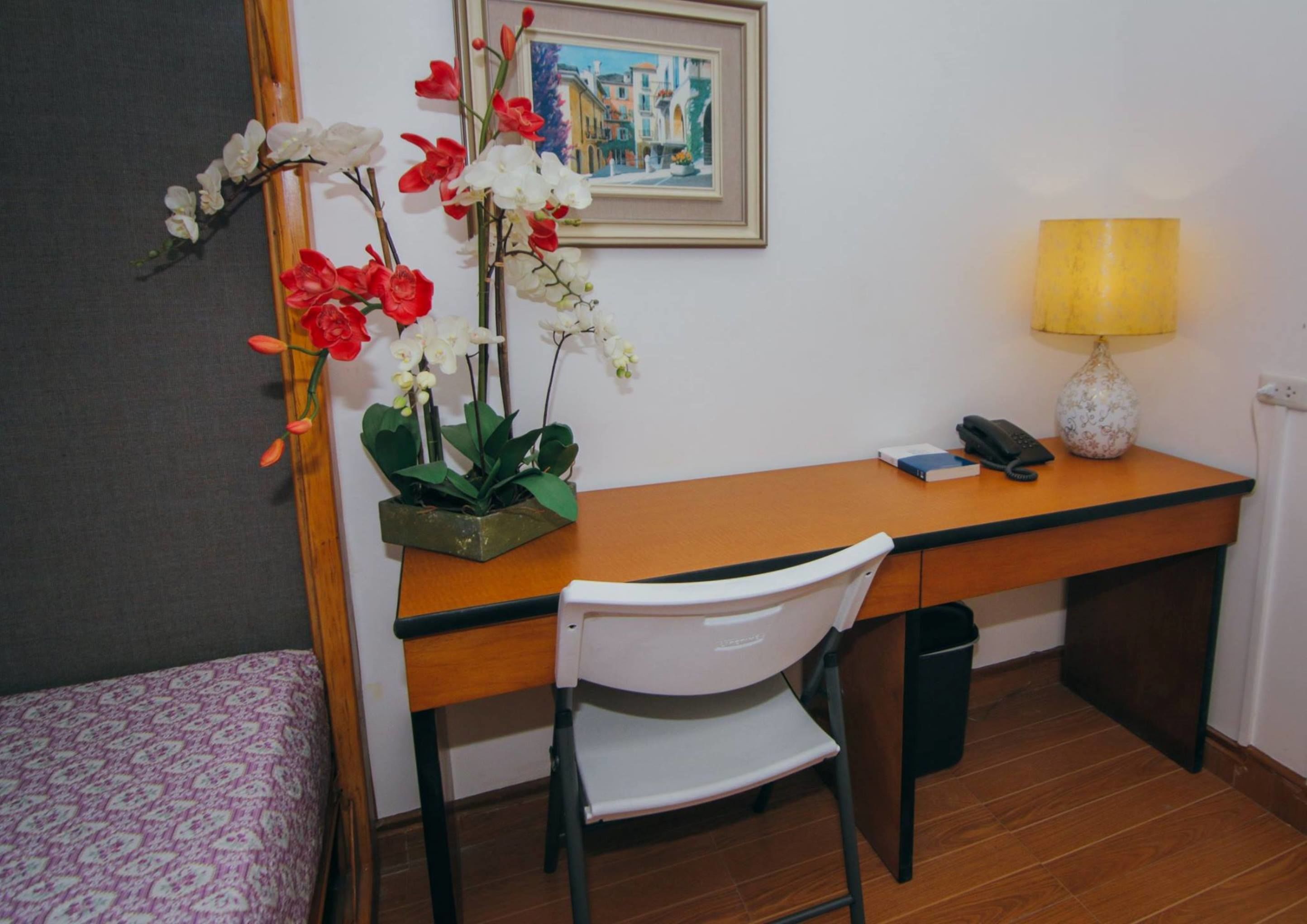 Quarto de hotel com uma escrivaninha de madeira, uma cadeira branca, uma pequena mesa de cabeceira com abajur, um quadro na parede e uma orquídea em vaso.