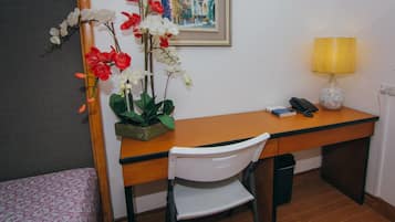 A hotel room with a wooden desk, a white chair, a small bedside table with a lamp, a framed picture on the wall, and a potted orchid plant.