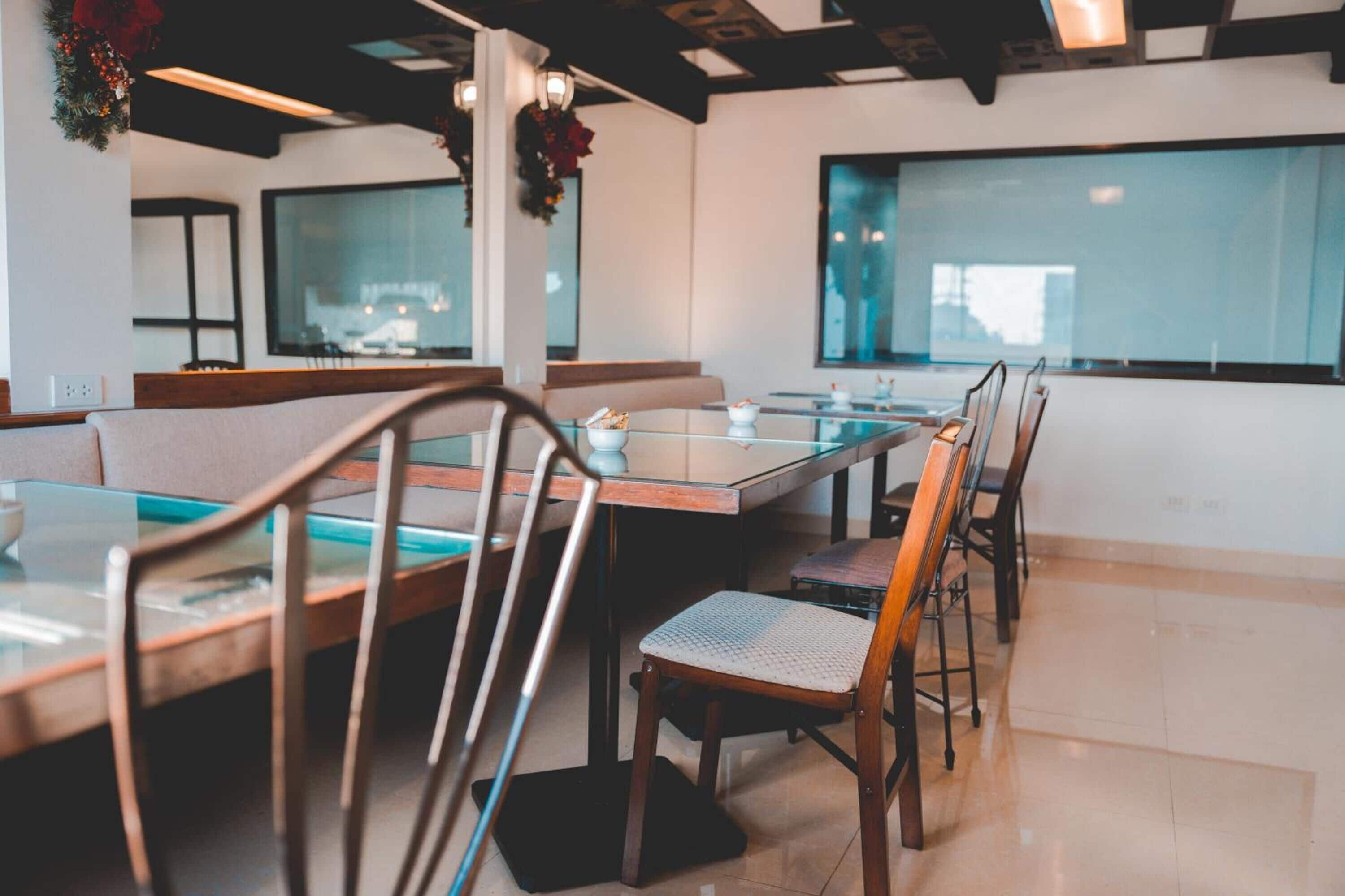Un restaurante con mesas de vidrio y sillas de madera, un techo alto con vigas a la vista y grandes ventanales.