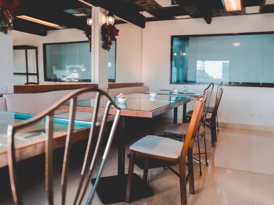 Een restaurant met glazen tafels, houten stoelen, een hoog plafond met zichtbare balken en grote ramen.