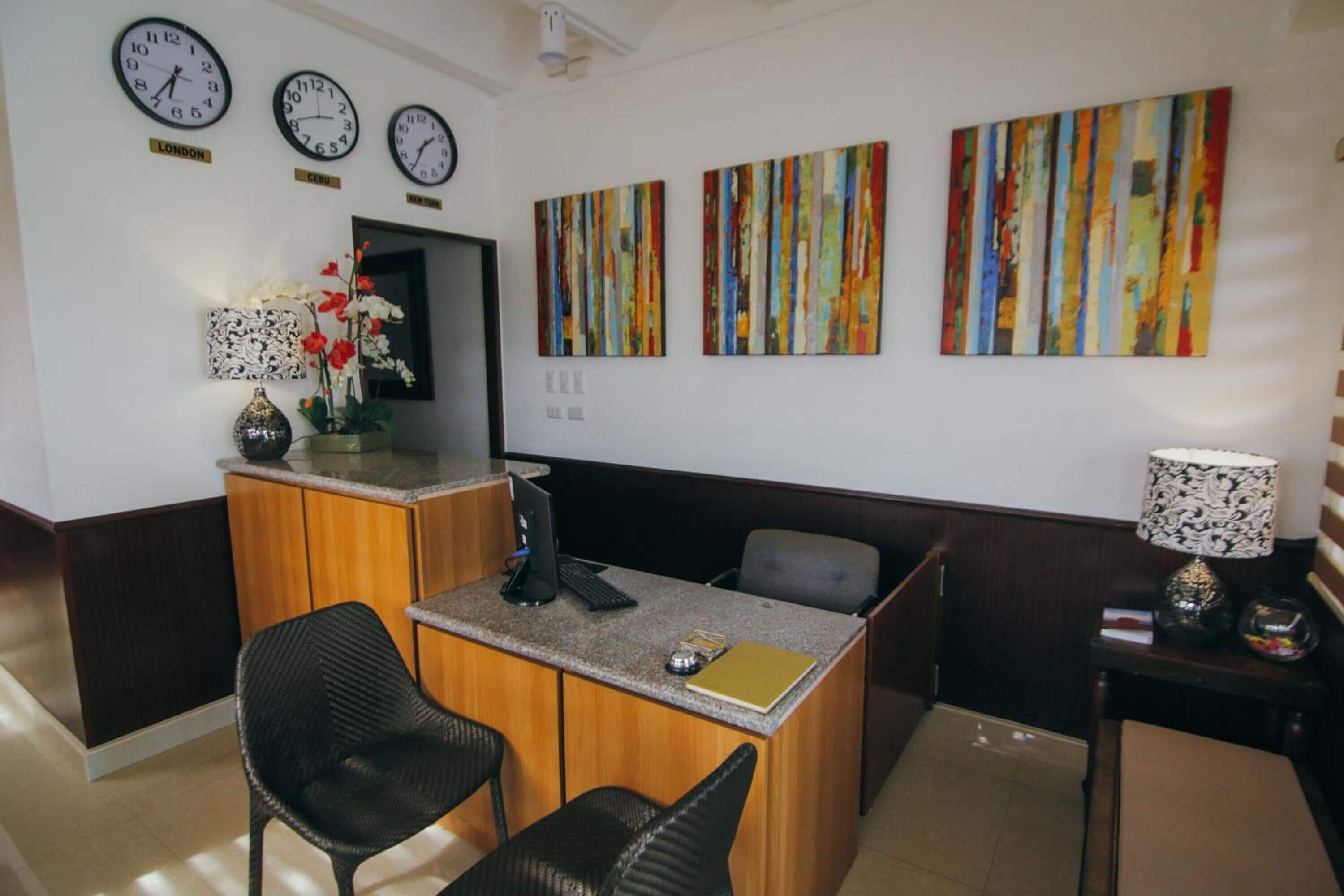 Un espace d’accueil comprenant un bureau, deux chaises, un vase avec des fleurs et trois horloges murales.