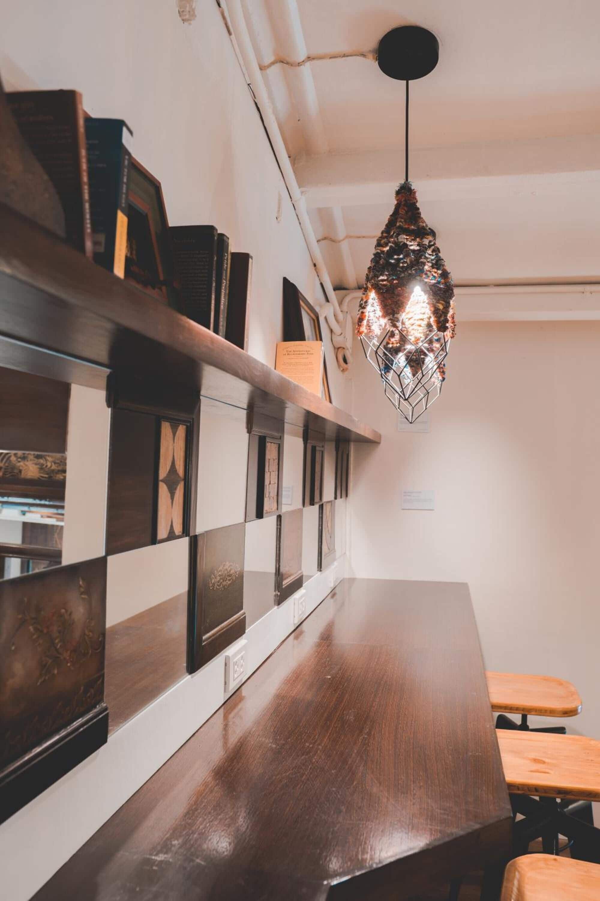 A wooden table with a metal frame, a hanging pendant light, and a wall with a checkered pattern and shelves with books.