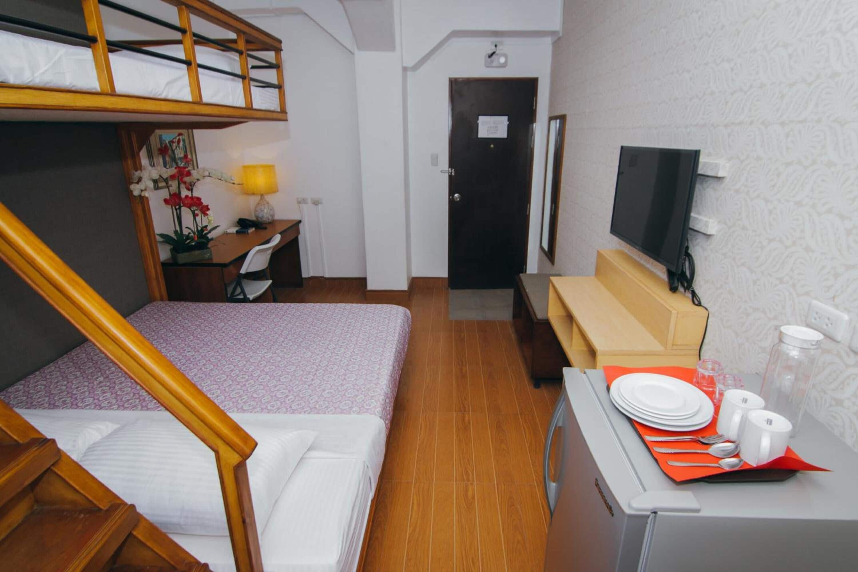 Una camera d'albergo compatta con un letto a castello, una scrivania con una lampada, una TV e un angolo cottura piccolo con un tavolo apparecchiato per mangiare.
