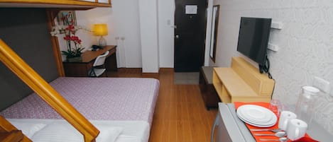 Une chambre d’hôtel compacte comprenant un lit superposé, un bureau avec une lampe, une télévision et une petite kitchenette avec une table dressée pour les repas.