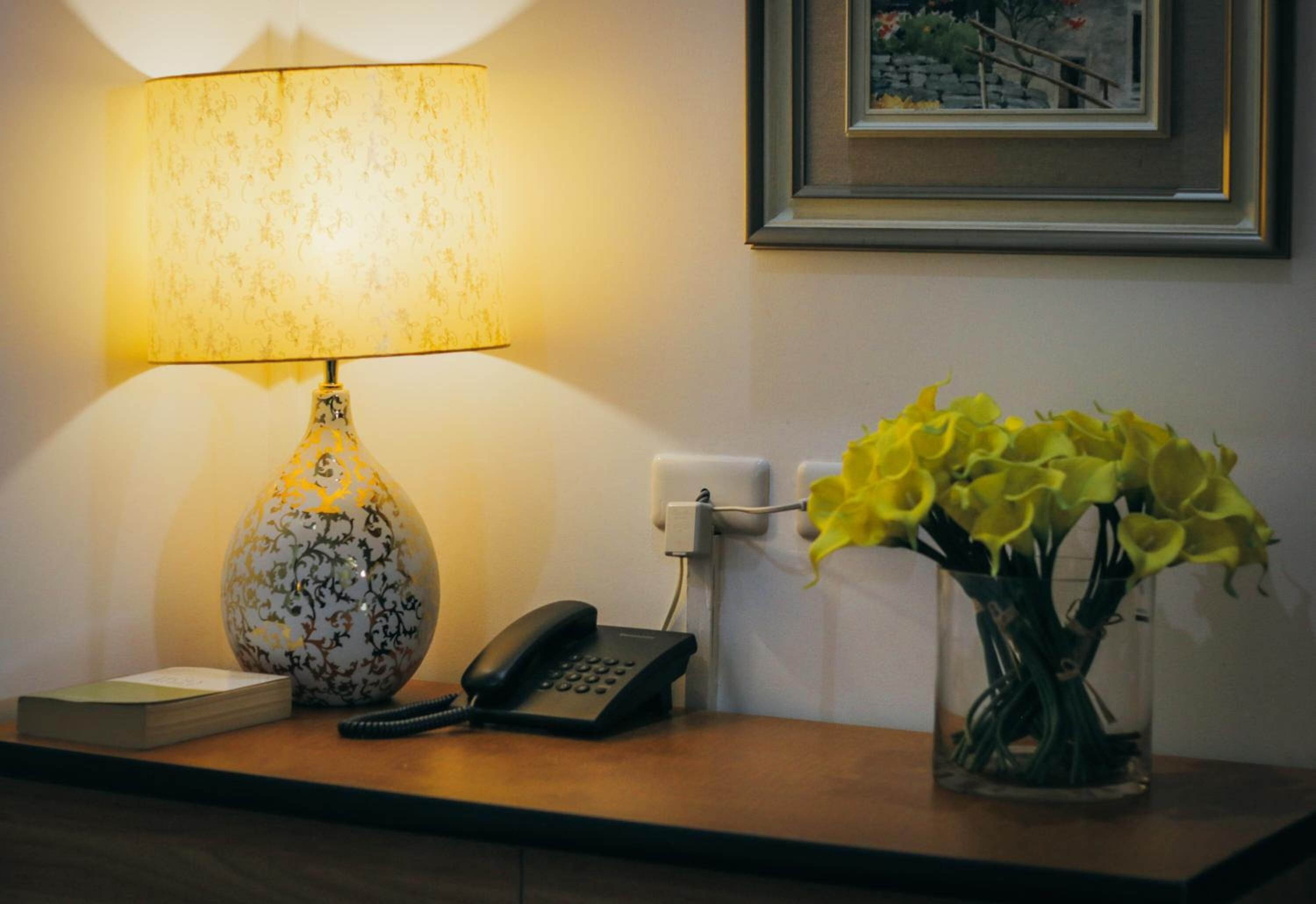 Un escritorio con una lámpara estampada, un teléfono, un florero con flores amarillas y un cuadro en la pared.