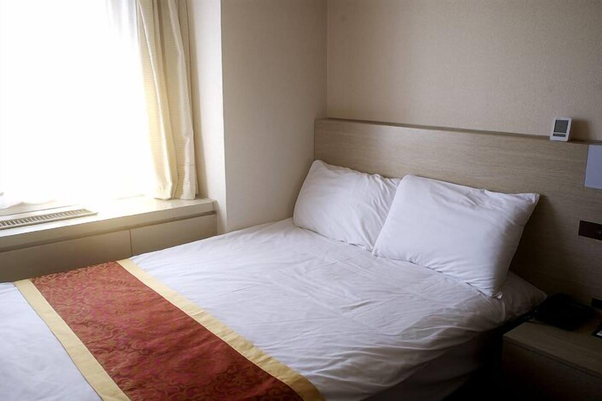 Una habitación de hotel con cama, mesita de noche, ventana con cortinas y un termostato en la pared.