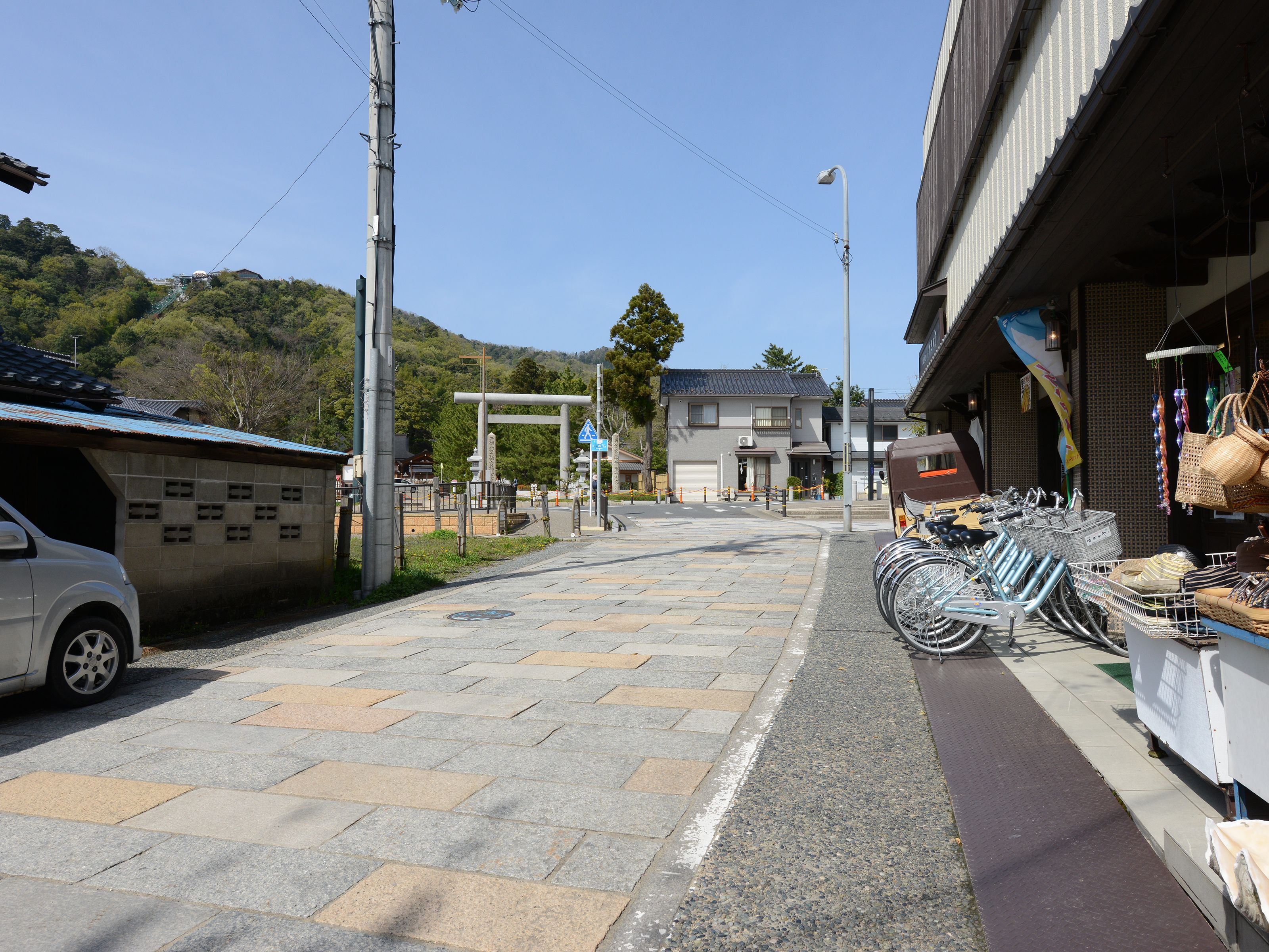 A street with a white car parked on the left, a row of bicycles on the right, and a building with a sign in the background.