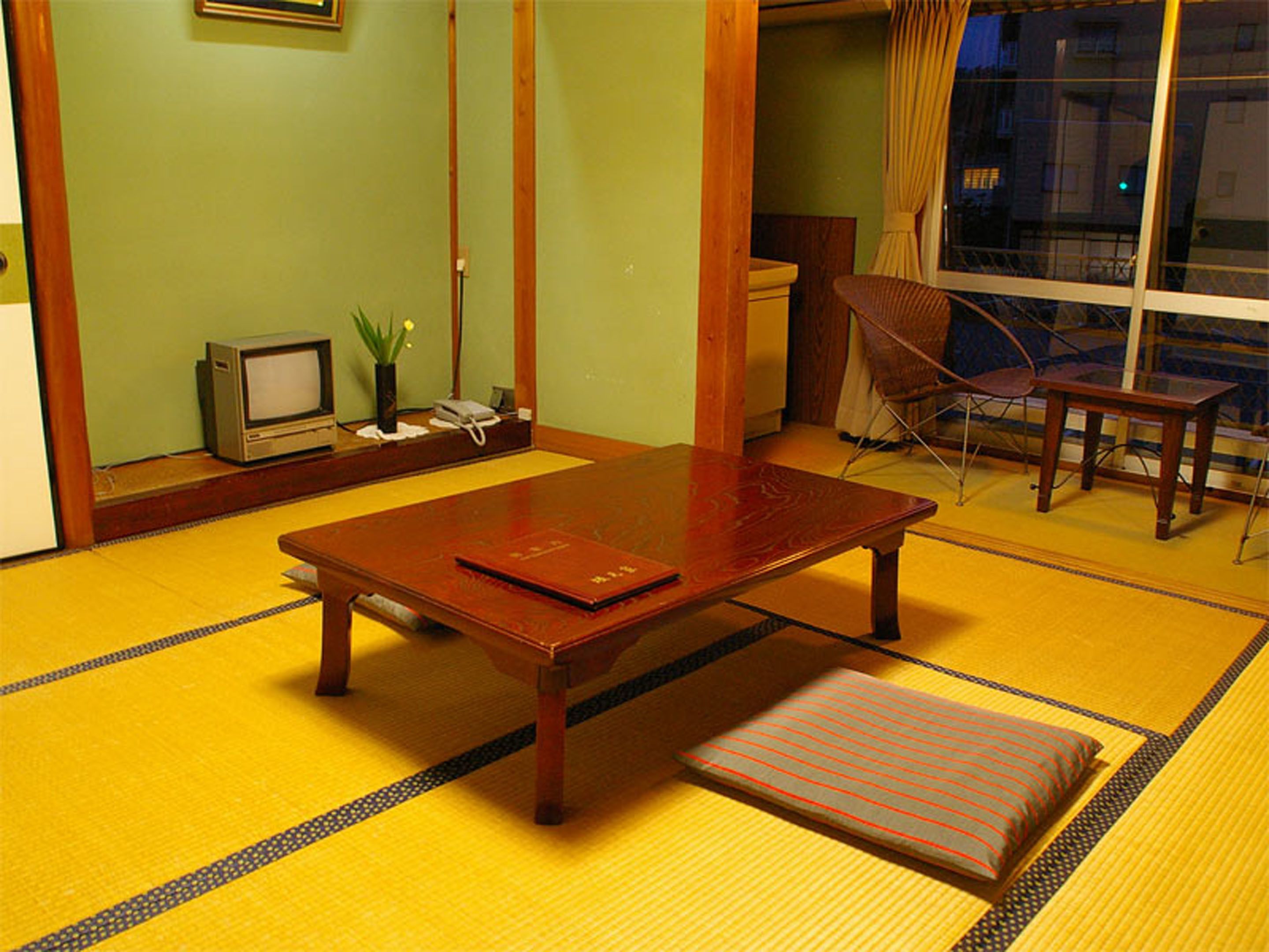 A traditional Japanese-style room with tatami flooring, a low wooden table, a cushioned bench, a television, and a window with curtains.
