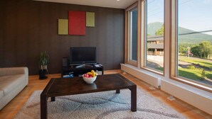 A modern living room with a dark wall, a flat-screen TV, a coffee table with fruit, and large windows offering a view of a building and mountains. - Holiday Niseko Shiki and Koyuki (Kutchan)