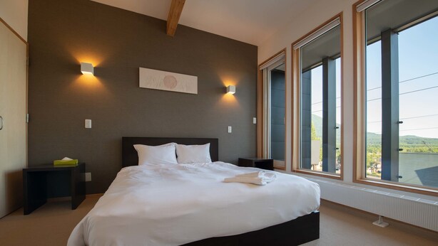 A neatly made bed in a room with large windows, a wall-mounted artwork, and two bedside lamps. - Holiday Niseko Shiki and Koyuki (Kutchan)