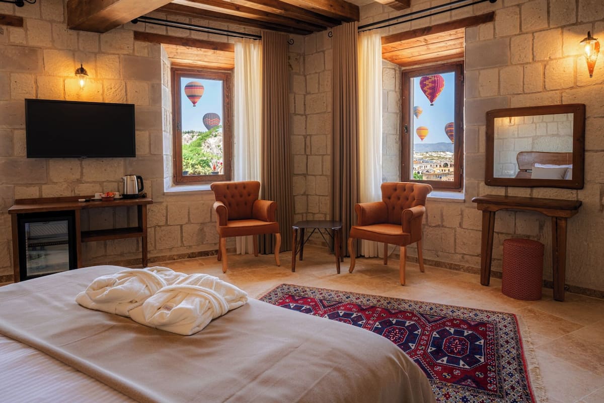 Una habitación de hotel acogedora con una cama grande, dos sillones, una mesita, un televisor y vistas a globos aerostáticos por las ventanas.