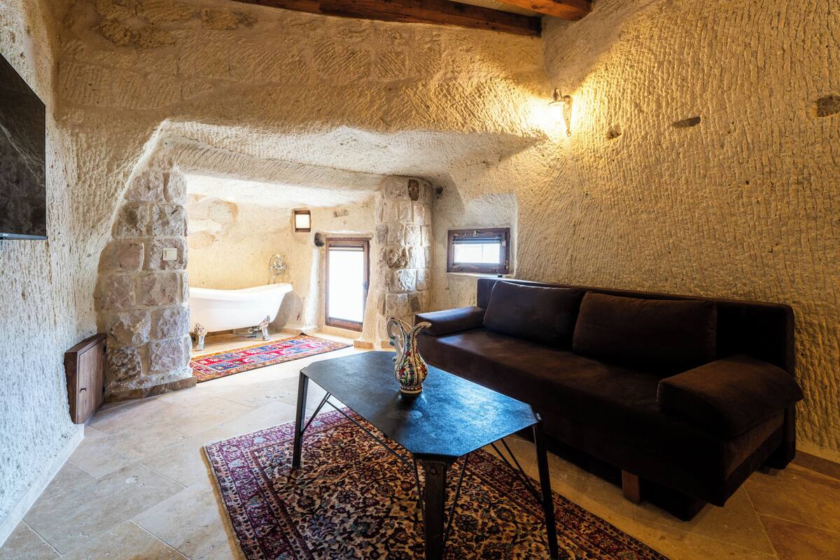 Una habitación con paredes de piedra, un sofá marrón oscuro, una pequeña mesa con un florero y una bañera al fondo.