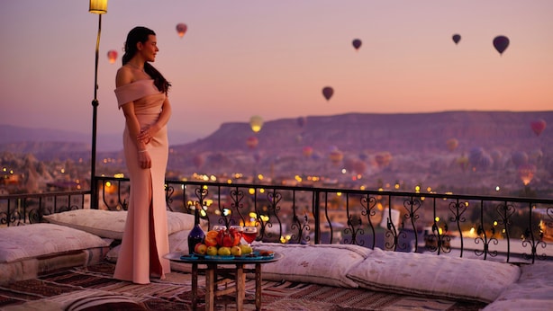 A balcony with a table set for two, overlooking a cityscape with hot air balloons. - Tulip Cave Suites (Nevsehir)