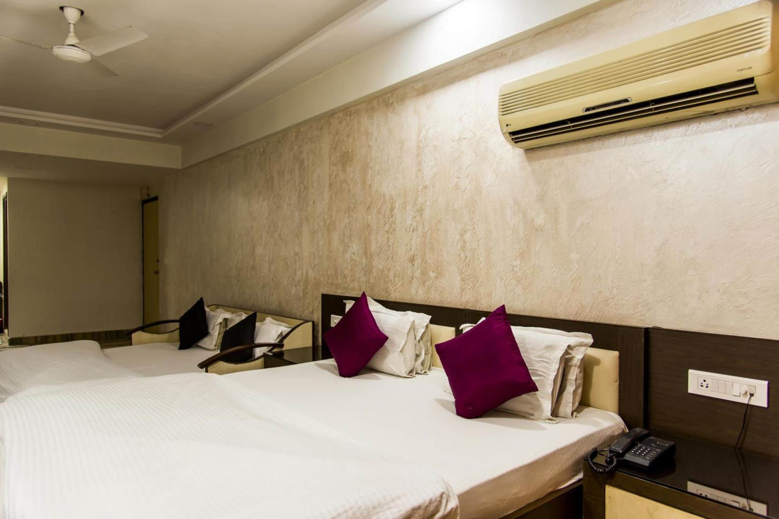 A hotel room with two beds, a ceiling fan, an air conditioning unit, a bedside telephone, and a wall-mounted electrical outlet.