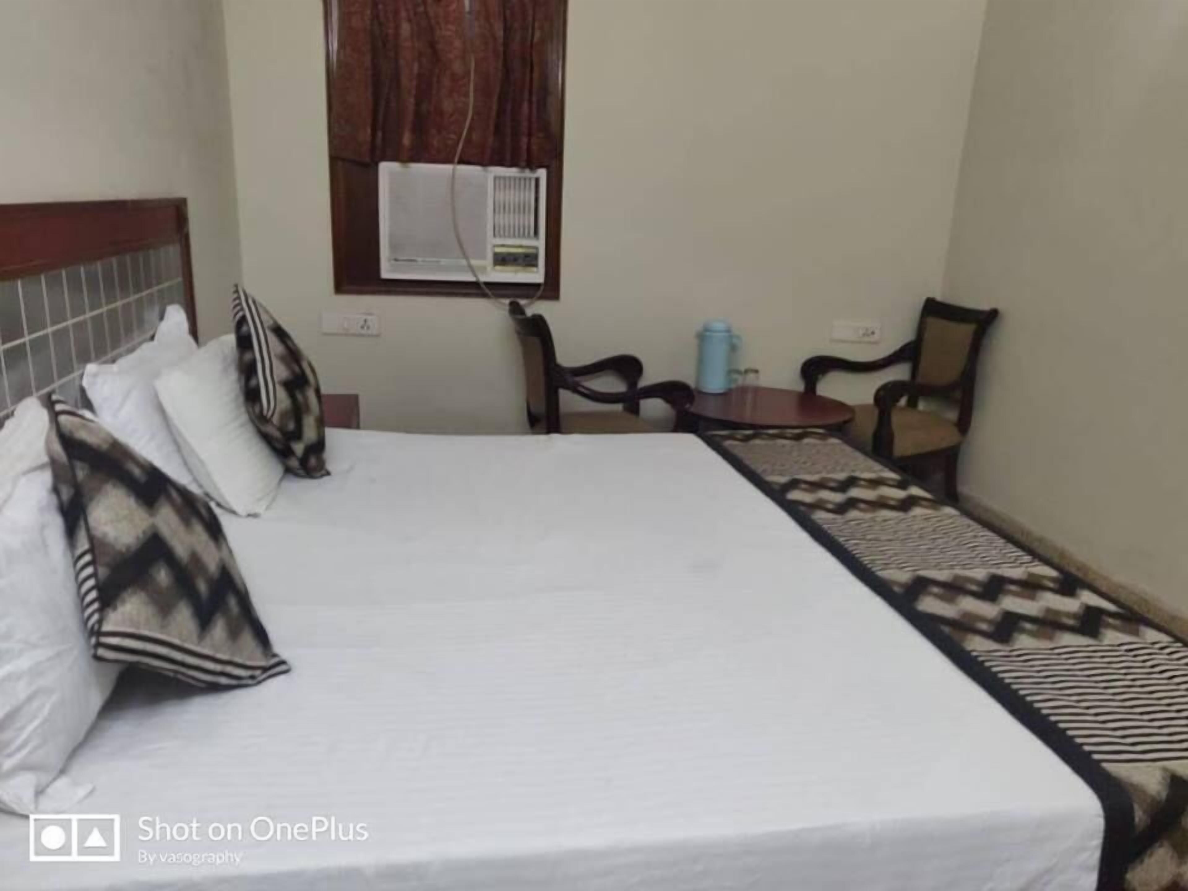 A hotel room with a bed, a window with curtains, a table with a chair, and a small table with a blue container.