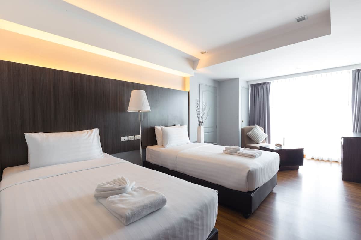 A hotel room with two beds, a bedside table with a lamp, a chair, and a large window with curtains.