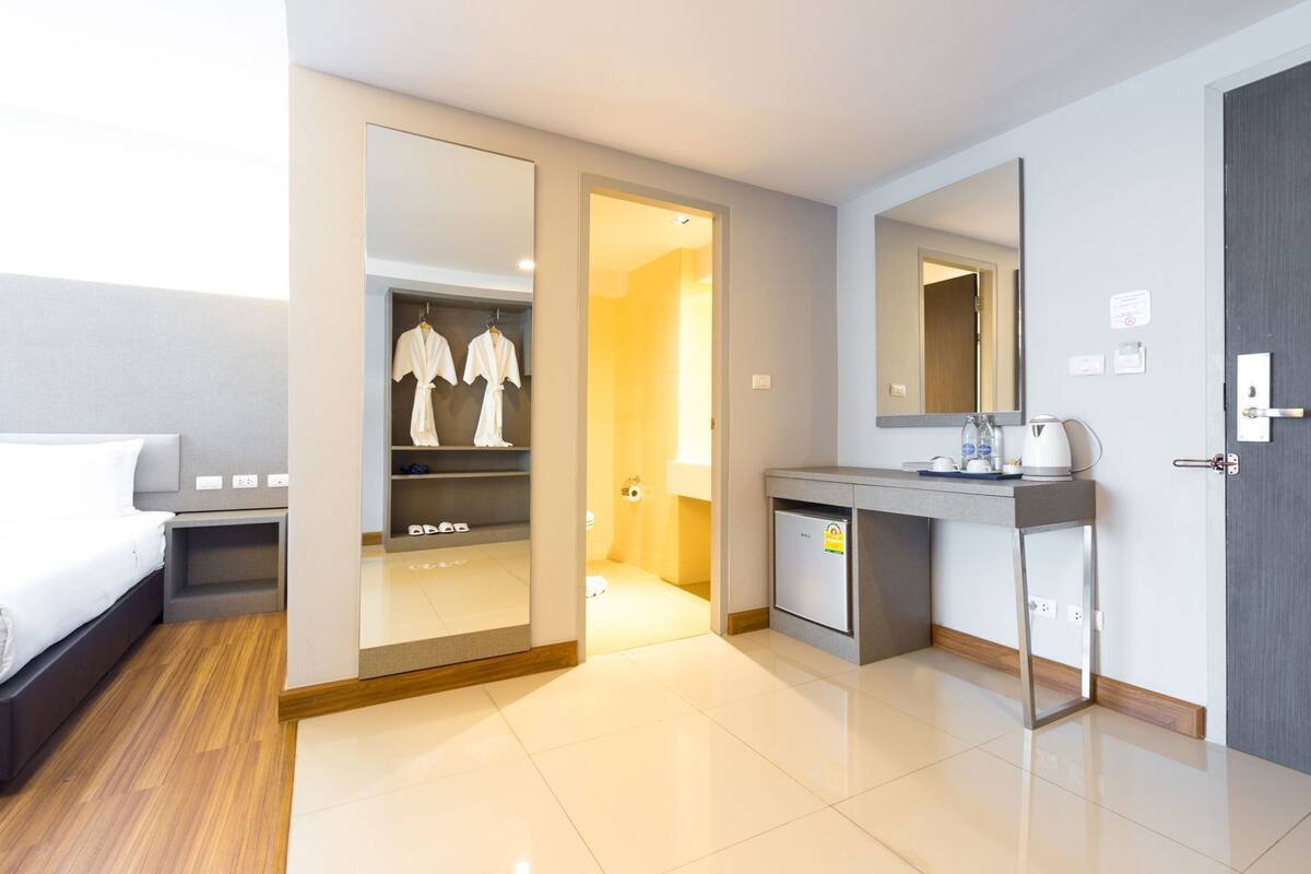 A modern hotel room with a bed, a walk-in closet with robes, a small kitchenette, and a large mirror.